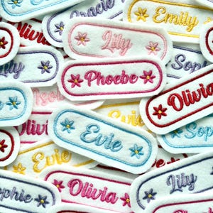 May include: A selection of embroidered name patches in different colours. Each patch displays a name in cursive script, like "Olivia" and "Evie", with a decorative border and floral details. The patches are white with colourful embroidery.