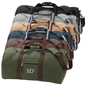 May include: A selection of canvas duffel bags in shades of black, grey, beige, navy, brown, and green. Each bag has a zipped main compartment, brown handles, and a detachable shoulder strap. The green bag is embroidered with the initials "SD".