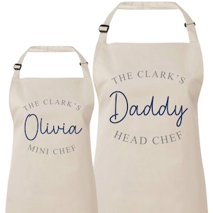May include: Two beige aprons with adjustable neck straps. One apron says "The Clark's Olivia Mini Chef" in navy blue and gray text. The other says "The Clark's Daddy Head Chef" in navy blue and gray text.