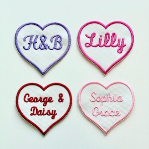 May include: Four heart-shaped embroidered patches. Each patch has a white centre with a different name or initials embroidered in a contrasting colour. The borders are purple, pink, red, and light pink.