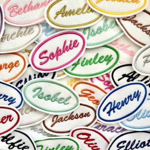 May include: A selection of embroidered name patches in different colours. Each patch displays a name in a cursive font, within an oval border. The patches feature shades of pink, green, blue, orange, and brown.