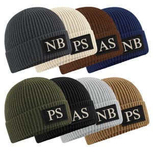 May include: A selection of ribbed knit beanies in shades like charcoal, cream, brown, navy, olive, black, grey, and tan. Each beanie has a rectangular patch with white embroidered initials. These beanies are a winter accessory.