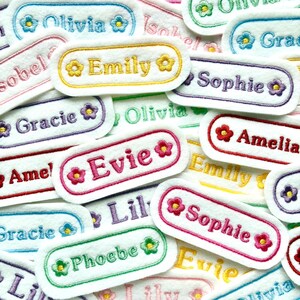 May include: A selection of embroidered name patches in various colours. Each patch features a name, such as "Emily" or "Sophie", in a contrasting colour, surrounded by a floral design. The patches are white with colourful borders.