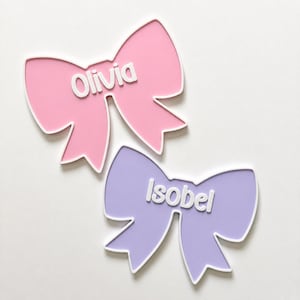 May include: Two bow-shaped wall decorations with white borders. The top bow is light pink with the white raised text Olivia. The bottom bow is light purple with the white raised text Isobel. Both items feature a smooth, flat surface design.