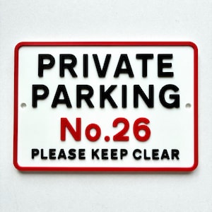 May include: A rectangular sign with a white background and a red border. The sign reads "PRIVATE PARKING No. 26 PLEASE KEEP CLEAR" in black and red lettering. The sign has two small holes for mounting.