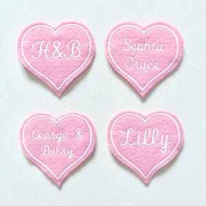 May include: Four pink heart-shaped patches with white embroidered borders and text. The patches feature the names "H&B", "Sophia Grace", "George & Daisy", and "Lilly" in white script. The hearts are arranged on a white surface.