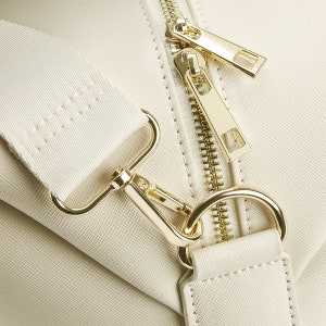 May include: Close-up of a cream-colored leather bag with a gold zipper and a gold hook and loop strap.