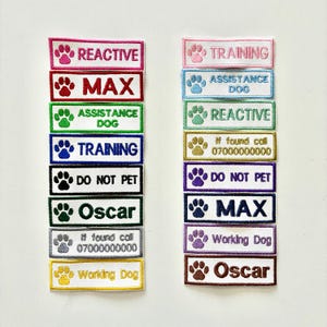 May include: A collection of embroidered dog patches in a range of colours, including pink, red, green, blue, black, and gold. Each patch displays text like "REACTIVE," "MAX," "TRAINING," and "DO NOT PET," alongside paw print motifs. These patches are for dogs.