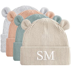 May include: A stack of knitted beanies in pastel colors: light gray, peach, teal, and cream. Each hat has matching ear-shaped accents. The frontmost cream-colored beanie features the embroidered letters "SM".