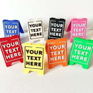 May include: A selection of small A-frame signs in various colours. Each sign displays the text "YOUR TEXT HERE" in a contrasting colour. The signs are in shades of blue, white, black, pink, red, yellow, orange, and green.