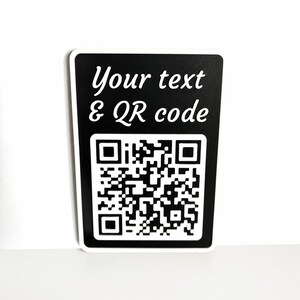 May include: A black rectangular sign with white text that reads "Your text & QR code." A large QR code is printed below the text. The sign has a white border and is set against a white background.