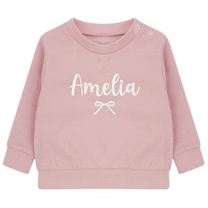 May include: A pale pink baby sweatshirt featuring the name "Amelia" in white script, with a bow detail. The long-sleeved top has a round neck and popper fastenings on the shoulder.