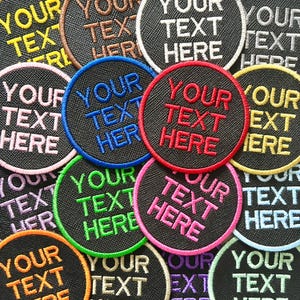 May include: A selection of circular embroidered patches, each with the text "YOUR TEXT HERE". The patches feature a black background and are bordered in various colours, such as blue, red, green, and pink. The text is in a contrasting colour to the border.