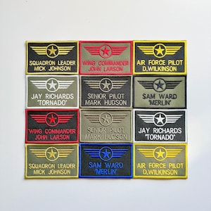 May include: A grid of twelve rectangular embroidered patches featuring aviation-themed wings and star emblems. Each patch displays a rank, a name, and a call sign in various colour combinations including black, olive green, red, blue, and yellow.