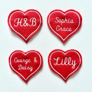 May include: Four red heart-shaped patches with white embroidered borders. Each heart features a different name or initials: "H&B", "Sophia Grace", "George & Daisy", and "Lilly". These patches are likely for crafts or personalization.