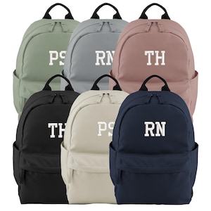 May include: Six backpacks in different colours: sage green, grey, dusty rose, black, cream, and navy blue. Each backpack has a white embroidered monogram. Features include a top handle, side pocket, and front pocket.