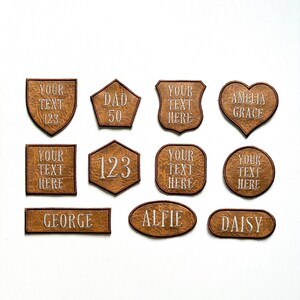 May include: A collection of eleven brown faux leather patches in various shapes including shields, hearts, circles, squares, and rectangles. Each patch features dark brown stitched borders and white embroidered text such as names and numbers.