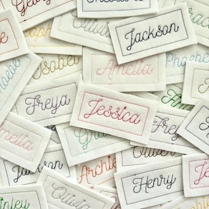 May include: A selection of embroidered name tags, each bearing a different name in cursive lettering. The tags are white with coloured stitching, such as red, blue, and green. Each tag is rectangular with a stitched border.