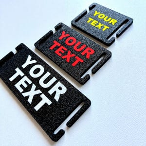May include: Three black rectangular tags with rounded corners and slots, each bearing the words "YOUR TEXT" in white, red, and yellow. The tags have a textured surface and are arranged diagonally.