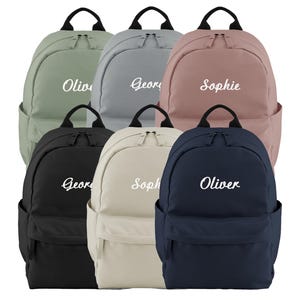 May include: Six backpacks in various colors: sage green, gray, dusty rose, black, cream, and navy blue. Each backpack has a front pocket, a top handle, and a name embroidered in white. The names are Oliver, George, and Sophie.