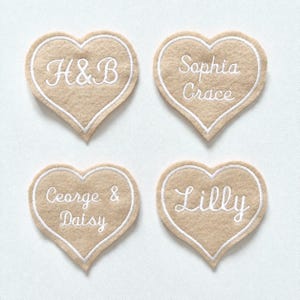 May include: Four beige felt heart-shaped patches with white embroidered text. The patches feature names and initials: "H&B", "Sophia Grace", "George & Daisy", and "Lilly". Each heart has a white border.