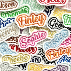 May include: A selection of embroidered name patches in diverse colours, such as orange, pink, blue, and gold. The names are in a cursive font with a contrasting border, creating a layered effect. The patches are on a white background.