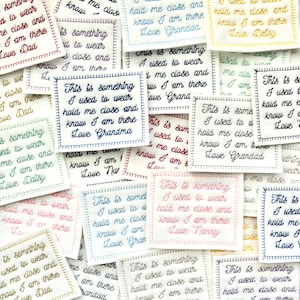 May include: A selection of embroidered fabric squares, each bearing the phrase "This is something I used to wear, hold me close and know I am there, Love [Name]" in different coloured threads. The squares are white.