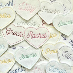 May include: A selection of white felt heart-shaped decorations, each embroidered with a name in different colours. Names such as "Lilly", "Rachel", "Isabelle", and "Gracie" are visible. The hearts have coloured stitching around the edges.