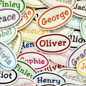 May include: A selection of embroidered name patches in assorted colours. Each patch displays a different name in a contrasting colour, including names such as Finley, George, and Oliver. The oval-shaped patches are made from a soft, felt-like material.