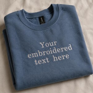 May include: A blue sweatshirt with white embroidered text reading "Your embroidered text here". The folded sweatshirt is made of a soft material. The brand tag is visible at the back of the neck.