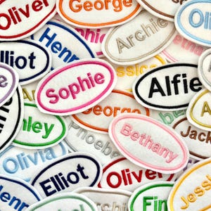 May include: A selection of embroidered name patches in various colours. Each patch is oval-shaped with a white background and a different name embroidered in a contrasting colour. Names include Sophie, Alfie, and Bethany.