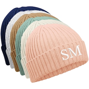 May include: A selection of ribbed knit beanies in shades of navy, grey, beige, teal, cream, and pink. The pink beanie has the embroidered initials "SM" in white. The beanies are displayed in a diagonal arrangement.