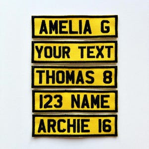May include: Five yellow rectangular patches with black text. The patches read: "AMELIA G", "YOUR TEXT", "THOMAS 8", "123 NAME", and "ARCHIE 16". The patches are likely for clothing or accessories.