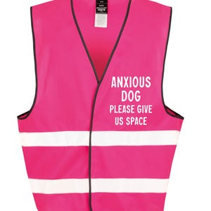 May include: A bright pink safety vest with black trim and two white reflective stripes. The vest features the text "ANXIOUS DOG PLEASE GIVE US SPACE" in white block letters, designed to alert others. The vest is designed for dogs.