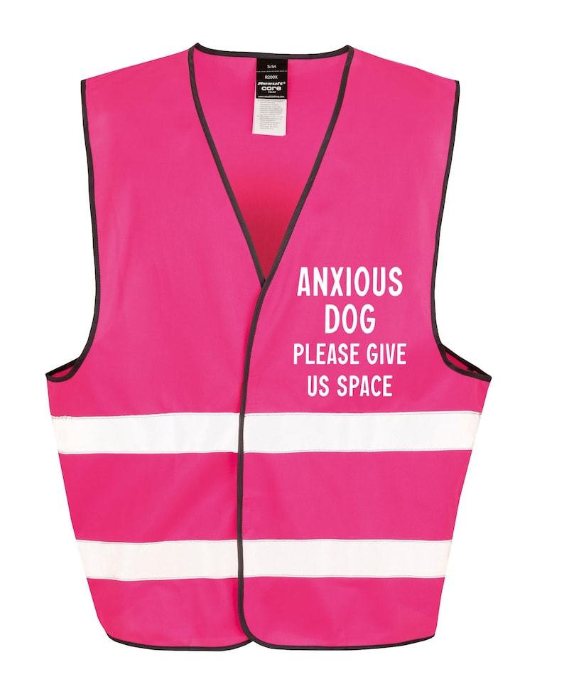 May include: A bright pink safety vest with black trim and two white reflective stripes. The vest features the text "ANXIOUS DOG PLEASE GIVE US SPACE" in white block letters, designed to alert others. The vest is designed for dogs.