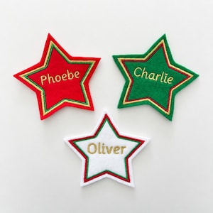 May include: Three star-shaped felt ornaments featuring embroidered names. One red star with green and gold trim reads Phoebe, one green star with red and gold trim reads Charlie, and one white star with red and green trim reads Oliver.