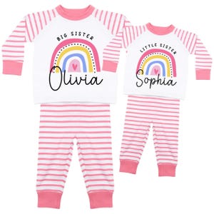 May include: Matching pink and white striped pajama sets. One set says "Big Sister" with a rainbow graphic and the name "Olivia". The other set says "Little Sister" with a rainbow graphic and the name "Sophia".