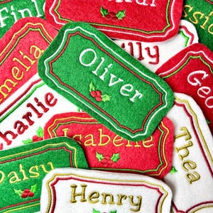 May include: A selection of Christmas name tags in red, green, and white. Each tag has a name embroidered in gold or white, with decorative borders and holly accents. The tags are made of felt.
