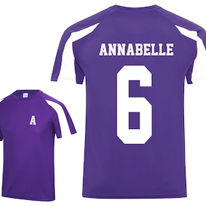 May include: Purple athletic shirt with white shoulder accents. The back of the shirt displays the name "ANNABELLE" and the number "6" in white block letters. A smaller shirt with the letter "A" is also shown.