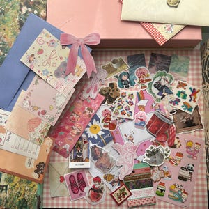 Pode incluir: Uma coleção de artigos de papelaria, incluindo autocolantes, cartões e envelopes. Os autocolantes apresentam vários desenhos, como personagens de desenhos animados, alimentos e padrões florais. Um laço de veludo rosa e uma caixa rosa também são visíveis.