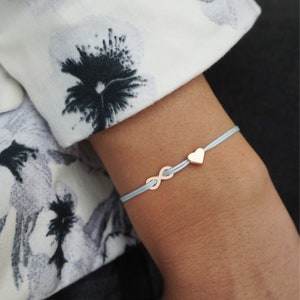 Könnte beinhalten: Ein hellblaues Kordelarmband mit einem kleinen Unendlichkeitssymbol in Roségold und einem passenden herzförmigen Anhänger in Roségold. Das Armband wird am Handgelenk getragen und ist vor einem weißen Stoffärmel mit schwarzem Blumenmuster zu sehen.