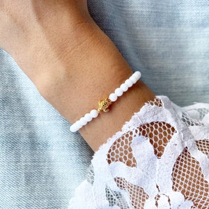 Könnte beinhalten: Weißes Perlenarmband mit einem goldenen Schildkrötenanhänger. Das Armband befindet sich am Handgelenk.