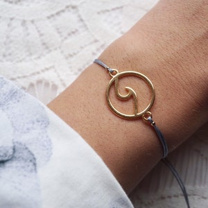 Könnte beinhalten: Ein goldfarbenes Armband mit einem kreisförmigen Design und einer wellenartigen Form in der Mitte. Das Armband ist an einer grauen Kordel befestigt. Das Armband befindet sich am Handgelenk einer Person, mit einem floral gemusterten Ärmel im Hintergrund.