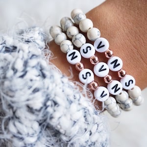 Könnte beinhalten: Drei Armbänder aus weißen Marmorperlen mit roségoldenen Abstandshaltern und schwarzen Buchstabenperlen, die den Namen "SVNS" buchstabieren.