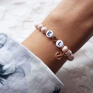 Könnte beinhalten: Ein Armband aus rosa und weißen Marmorperlen mit goldenen Akzenten. Das Armband trägt die Buchstaben "E" und "L" sowie einen kleinen goldenen Anker-Anhänger.