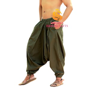 May include: A man wearing a pair of olive green loose-fitting pants with a drawstring waist. The pants have a large pocket on the right side. The man is standing with his right hand in the pocket. The pants are made of a soft, comfortable fabric.  "Sarjana Handicrafts" is printed on the pants.