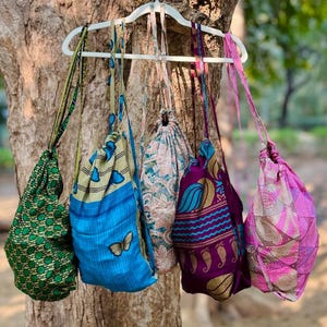 May include: Five drawstring bags of various colors and patterns hang from a white hanger. The bags are made of fabric with designs in green, blue, pink, and purple. One bag has a butterfly applique.