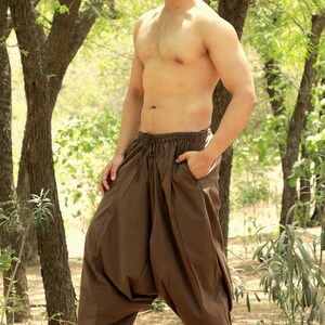May include: A man wearing brown loose-fitting pants with a drawstring waist. He is standing in a forest setting with trees and green foliage in the background.