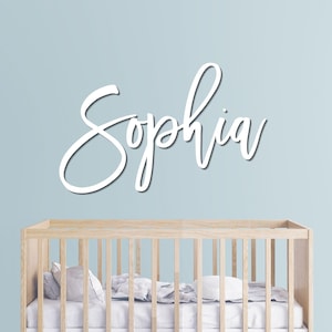 May include: White cursive wooden sign that reads "Sophia" against a light blue wall. A wooden crib is in the foreground, with pillows and bedding visible inside. The sign is a decorative element, likely for a nursery or child's room.