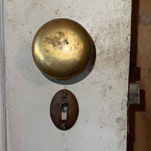 May include: A round, tarnished brass doorknob mounted on a white painted wooden door. Below the knob is an oval-shaped, dark metal keyhole escutcheon plate secured with two small screws, featuring a vertical slot for a key mechanism.